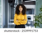 Confident businesswoman with curly hair in yellow blouse stands with arms crossed in modern office hallway. Strong, accomplished professional representing leadership, success, empowerment.