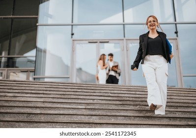 Confident businesswoman descending stairs of a modern office building, holding documents and phone, with colleagues interacting in the background. - Powered by Shutterstock