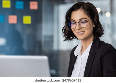 Confident businesswoman with glasses smiling while working on laptop in a modern office environment. Professional and focused on her tasks. - Powered by Shutterstock