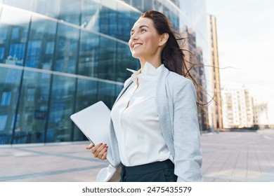 Confident businesswoman, holding a laptop, stands smiling against a backdrop of modern city architecture.

 - Powered by Shutterstock