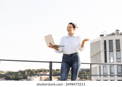 Confident businesswoman holding laptop, working on rooftop, smiling happily. Casual attire, outdoor setting. High-rise building in background, clear skies. - Powered by Shutterstock