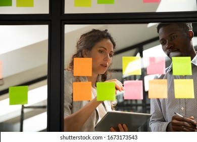 Confident businesswoman mentor organizing work, explaining project plan, tasks on stickers to African American trainee, diverse employees working together, discussing strategy, sharing startup ideas