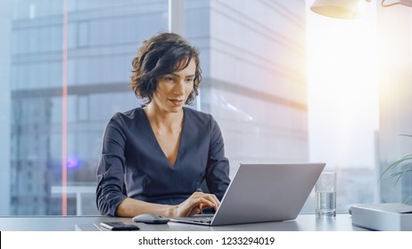 Confident Businesswoman Sitting At Her Desk And Working On A Laptop In Her Modern Office. Stylish Beautiful Woman Doing Important Job. In The Window Big City Business District View With Sun Flare.