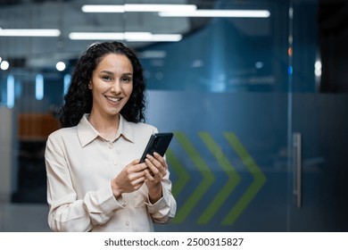 Confident businesswoman smiling at camera while using phone in modern office. Professional attire, relaxed atmosphere, tech-savvy individual. - Powered by Shutterstock