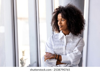 A confident businesswoman stands by a large window, gazing thoughtfully outside. She exudes professionalism and determination in a bright white shirt, embodying modern office style and ambition. - Powered by Shutterstock