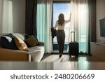 Confident businesswoman wearing white shirt and holding suit with suitcase stands in a hotel room while looking at the view from window Beautiful sunset. Woman enjoying her holiday in a luxury hotel.