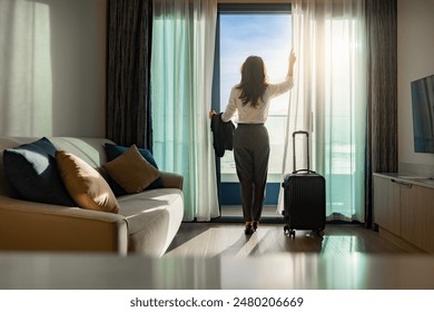 Confident businesswoman wearing white shirt and holding suit with suitcase stands in a hotel room while looking at the view from window Beautiful sunset. Woman enjoying her holiday in a luxury hotel. - Powered by Shutterstock