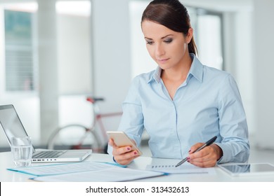 Confident businesswoman working at office desk and using a touch screen smart phone, room interior on background