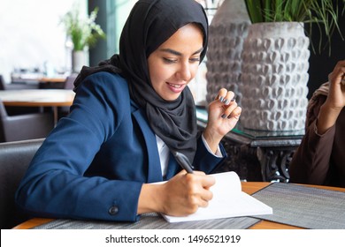 Confident businesswoman working on business plan in coffee shop. Young Muslim business woman in hijab and office suit writing notes in cafe. Work in cafe concept - Powered by Shutterstock