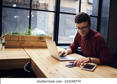 Confident busy male freelancer in trendy eyewear doing remote job using modern technologies checking installation of application on smartphone while surfing net searching information for presentation