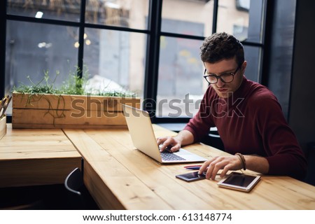 Confident busy male freelancer in trendy eyewear doing remote job using modern technologies checking installation of application on smartphone while surfing net searching information for presentation