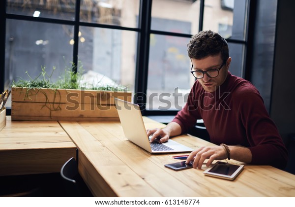Confident busy male freelancer in trendy\
eyewear doing remote job using modern technologies checking\
installation of application on smartphone while surfing net\
searching information for\
presentation