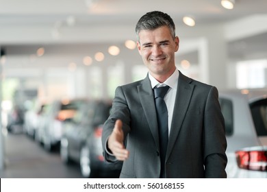 Confident car dealer giving a handshake, car showroom on the background