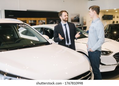 Confident Car Salesman Wearing Business Suit Is Telling About New Car Model To Young Man Wearing Casual Clothes In Auto Dealership. Concept Of Choosing And Buying New Car At Showroom.