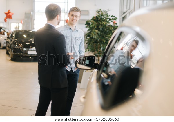 Confident car\
salesman wearing business suit is telling about new car model to\
young man wearing casual clothes in auto dealership. Concept of\
choosing and buying new car at\
showroom.