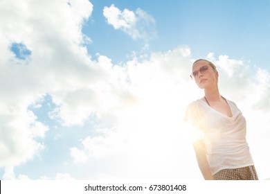 Confident Casual Woman Wearing White T-shirt And Red Sunglasses Relaxing In Nature Shot From Below Against Bright Summer Sky With Sun Rays.