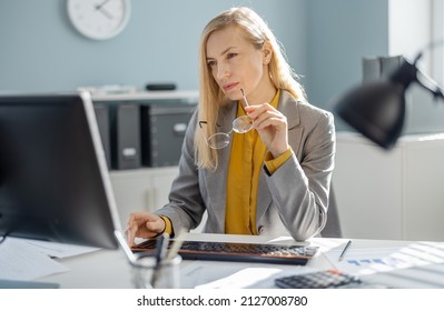 Confident Caucasian Business Woman In Formal Suit Typing On Modern Pc While Working At Office. Mature Lady With Blond Hair Sitting At Desk And Looking On Monitor With Focused Facial Expression.