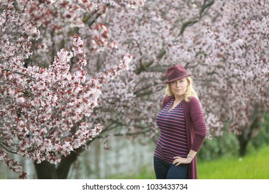 Confident Caucasian Woman With Long Blonde Hair Near Blossoming Orchard With Purple Fedora Hat