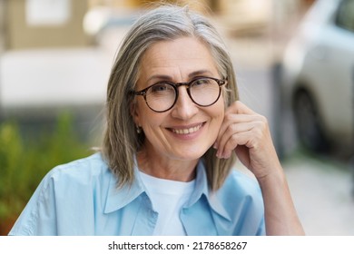 Confident Charming Mature Woman Smiling Leaned Head On Hand Wearing Eye Glasses And Blue Shit Sitting Outdoors With City Background. Portrait Of Mature Woman With Grey Hair And Good Skin.