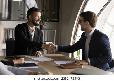 Confident cheerful boss and project manager man in formal suits shaking hands over meeting table, smiling, discussing deal, contract, investment, project success, starting partnership, cooperation - Shutterstock ID 2392473593