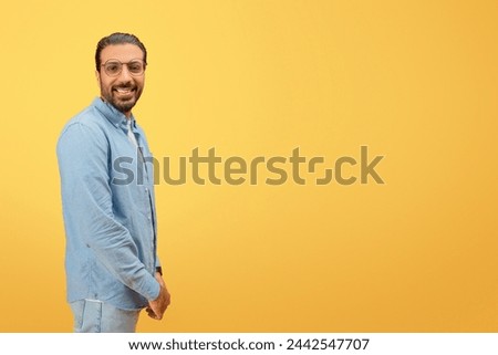 Confident and cheerful indian man with glasses and beard posing against a yellow background