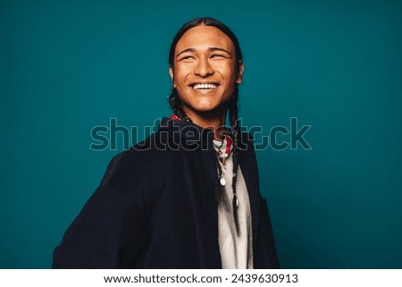 Confident, cheerful and stylish young man with braided hair stands against a blue background. Ethnic jewelry and casual clothing complement his unique identity. 
