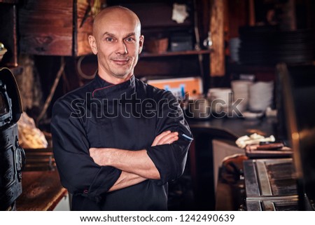 Confident chef posing with his arms crossed and looking at a camera in restaurant kitchen.