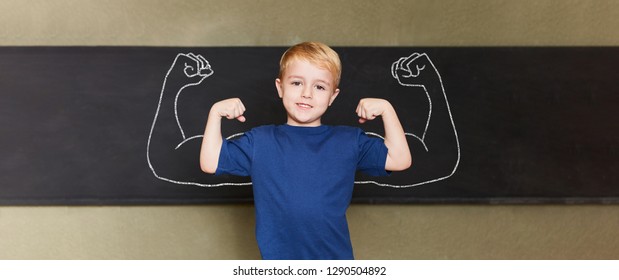 Confident child stands with muscles in front of the blackboard in a primary school - Shutterstock ID 1290504892