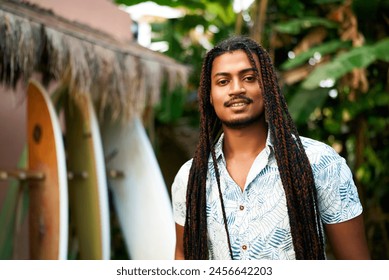 Confident coach with dreadlocks stands at surf camp. Surfboards align in background. Instructor with warm smile invites for lessons. Black surfer promotes sport, inclusion tropical retreat. - Powered by Shutterstock