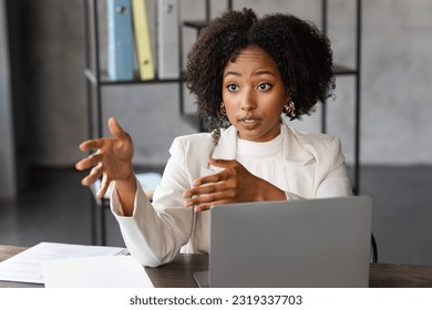 Confident corporate beautiful young african american businesswoman in white formal outwear sitting at desk in front of laptop, talking and gesturing, have interview at office, copy space - Powered by Shutterstock