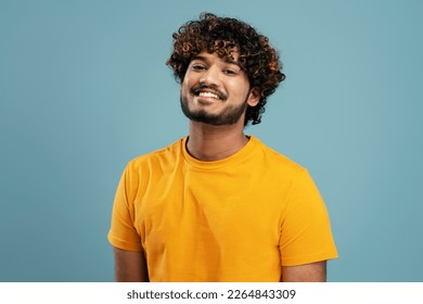 Confident  curly haired bearded Indian man, wearing a bright yellow t-shirt, smiling a beautiful toothy smile looking at camera, isolated over blue background - Powered by Shutterstock