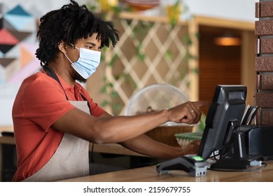 Confident Curly Millennial Black Male Bartender In Apron, Protective Mask At Bar Counter With Terminal, Works On R-keeper In Cafe Interior. Money For Services, Finances And Small Business, Covid-19