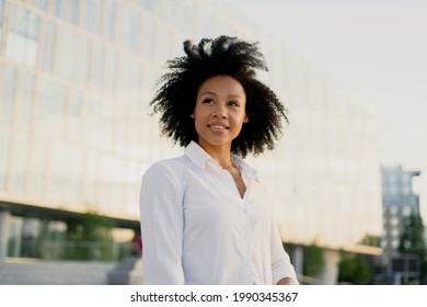 A Confident, Curly-haired Young Woman Of African-American Appearance Stands Outside A Business Center In The City And Smiles. Dressed In A White Shirt. A Confident Person On The Street.