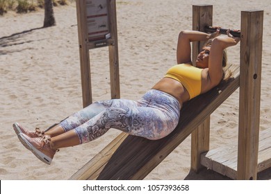 Confident Curvy Blond Woman In Sportswear Doing Abs Exercise On Wooden Plank With Bar On Sandy Beach In Sunlight.