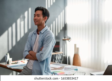 Confident Designer. Side View Of A Young Asian Man In Casual Wear With Crossed Arms Looking Away And Smiling While Standing In The Modern Office. Creative People