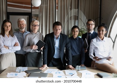 Confident Diverse Employees Of Different Age Standing At Meeting Table Together, Looking At Camera. Multiethnic Young And Senior Interns, Students, Mentor, Teacher Team Portrait. Corporate Head Shot