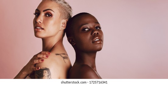Confident diverse female models standing back to back and looking at camera. Two multi-ethnic women standing together over beige background.