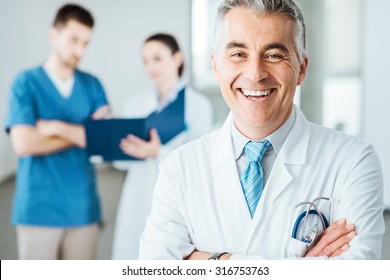 Confident doctor posing and smiling at camera and medical staff checking medical records on background