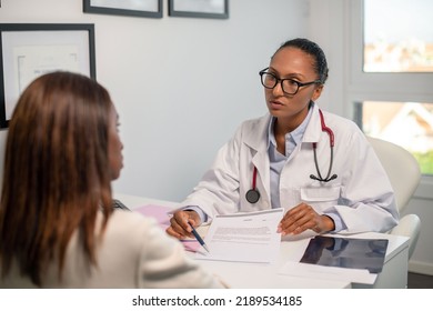 Confident Doctor Showing Agreement To Female Patient In Clinic. Young African American General Practitioner Wearing Glasses Giving Document To Woman. Medical Services Contract Concept