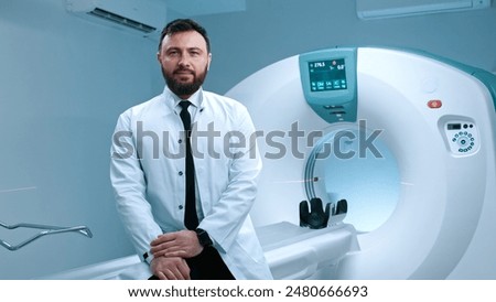 Confident doctor sitting at capsule of magnetic resonance imaging. Bearded doctor in glasses. Modern equipped room for MRI scanning. Doctor in white gown at background of MRI capsule.