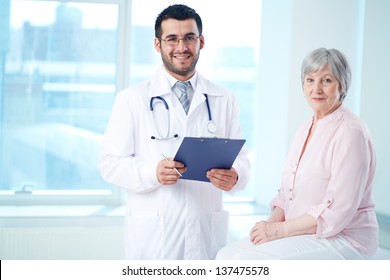 Confident Doctor With Stethoscope And Clipboard And His Senior Patient Looking At Camera