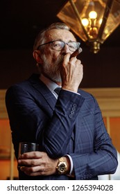 Confident Elderly Man In Blue Expensive Suit Clothes Enjoying A Rest, Relaxing In Private Gentleman's Club, Sipping Old Whiskey And Smoking Cigars Is The Way He Relaxes.