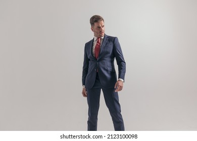 confident elegant man in suit with red tie looking to side, being cool and standing in a powerful pose on grey background in studio