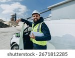 Confident engineer holding blueprint while standing by his van at a concrete production plant, symbolizing industrial development and construction