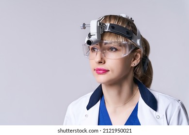 Confident ENT Doctor Wearing Surgical Headlight Head Light And Protective Glasses. Closeup Portrait Of Female Otolaryngologist Or Head And Neck Surgeon On Light Grey Background.