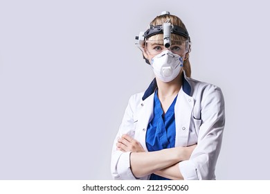 Confident ENT Doctor Wearing Surgical Headlight Head Lantern, Protective Face Mask And Glasses. Portrait Of Female Otolaryngologist Standing Arms Crossed On Light Grey Background.