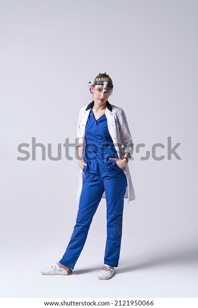 Confident ENT\
doctor wearing surgical headlight head light and protective\
glasses. Full length portrait of female otolaryngologist or head\
and neck surgeon on light grey\
background.