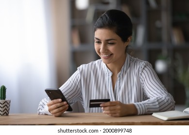 Confident ethnic female student pay tuition fee remotely using mobile phone bank app. Smiling young indian lady sit at office table use credit card to make online money transfer buy goods at internet