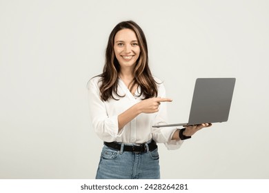 Confident european woman holding and pointing at a laptop screen, dressed in a white blouse and blue jeans, symbolizing ease of use in technology and education, studio. Business, work - Powered by Shutterstock