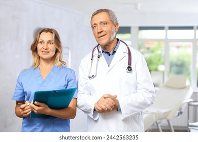 Confident experienced elderly male doctor in white coat with phonendoscope around neck standing with female assistant in blue scrub holding folder with patient history - Powered by Shutterstock